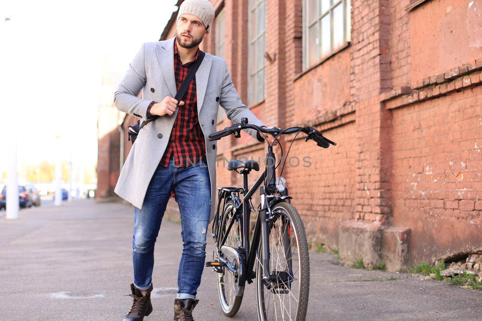 Handsome young man in grey coat and hat pushing his bike down a street in the city
