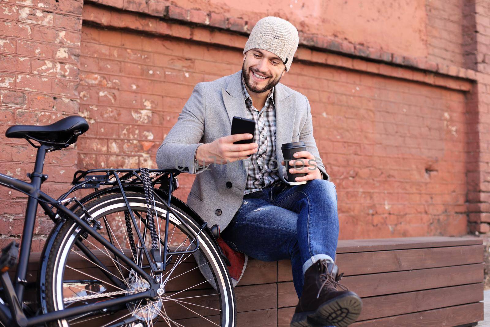Handsome young man in grey coat and hat talking on the mobile phone and smiling while sitting near his bicycle outdoors