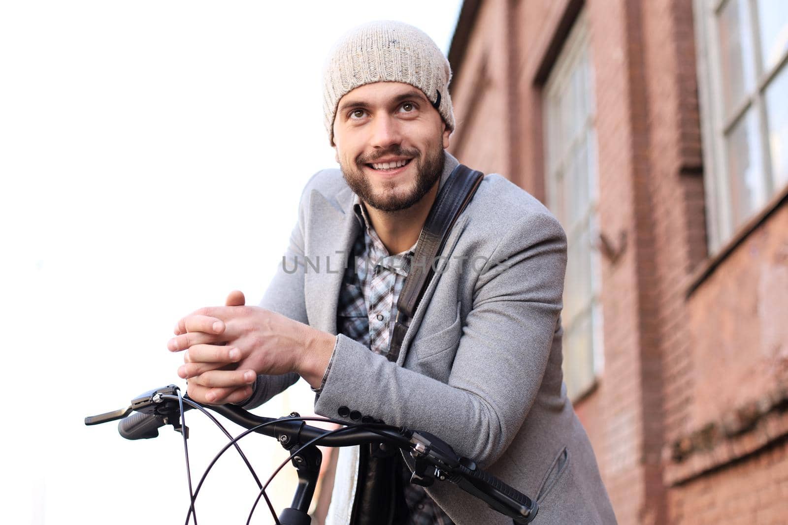 Handsome young man in grey coat and hat standing with his bicycle