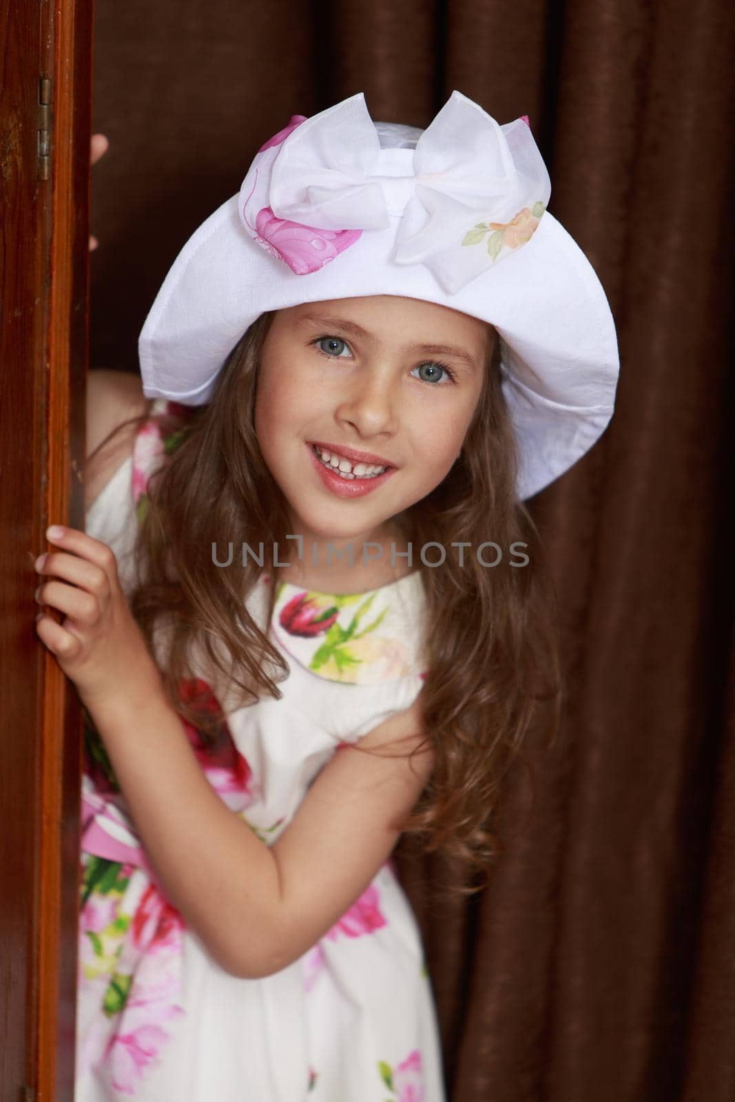 Cheerful little girl in a long dress and a hat peeks out from behind the wardrobe