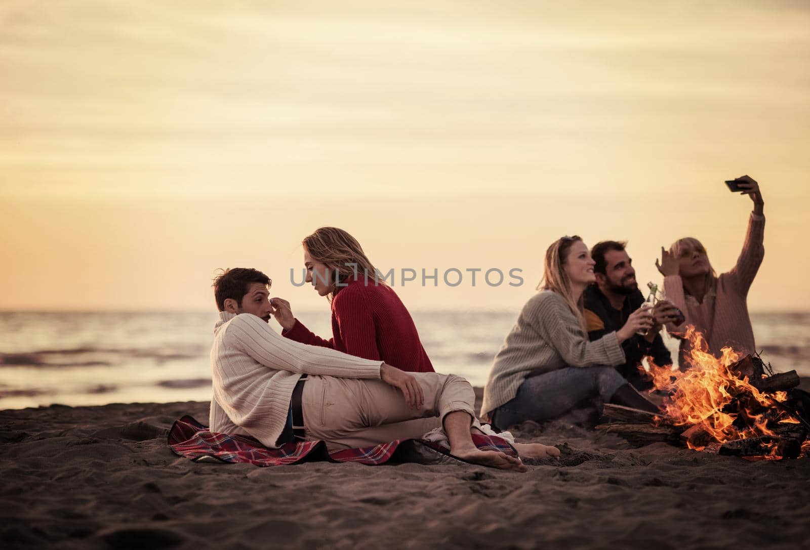 Happy Carefree Young Friends Having Fun And Drinking Beer By Bonefire On The Beach As The Sun Begins To Set filter