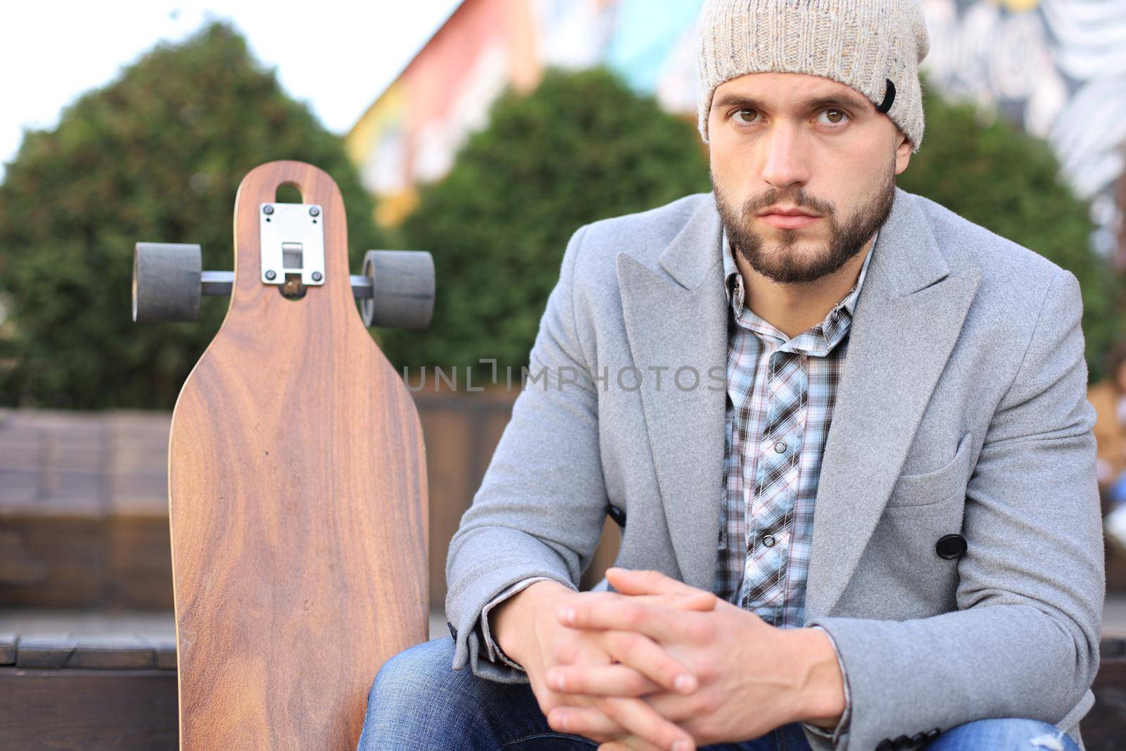 Handsome young man in grey coat and hat, resting, sitting with longboard. Urban skateboarding concept