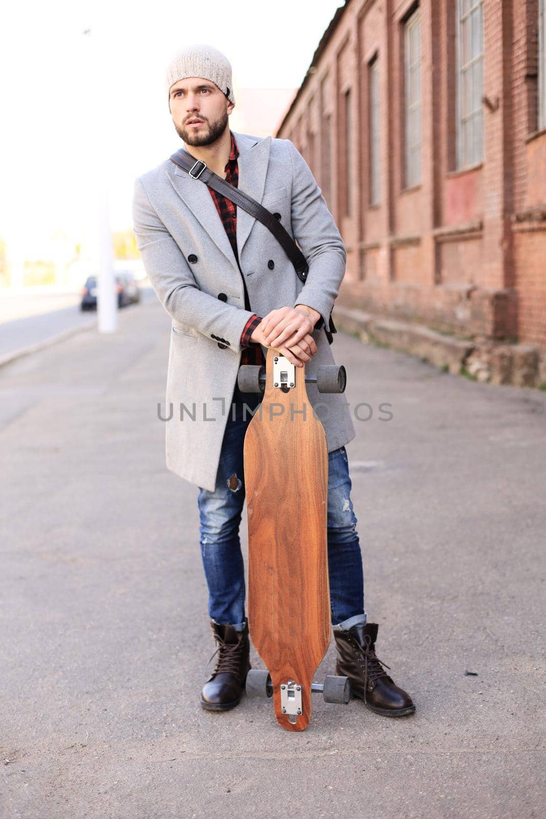 Handsome young man in grey coat and hat walking on the street, using longboard. by tsyhun