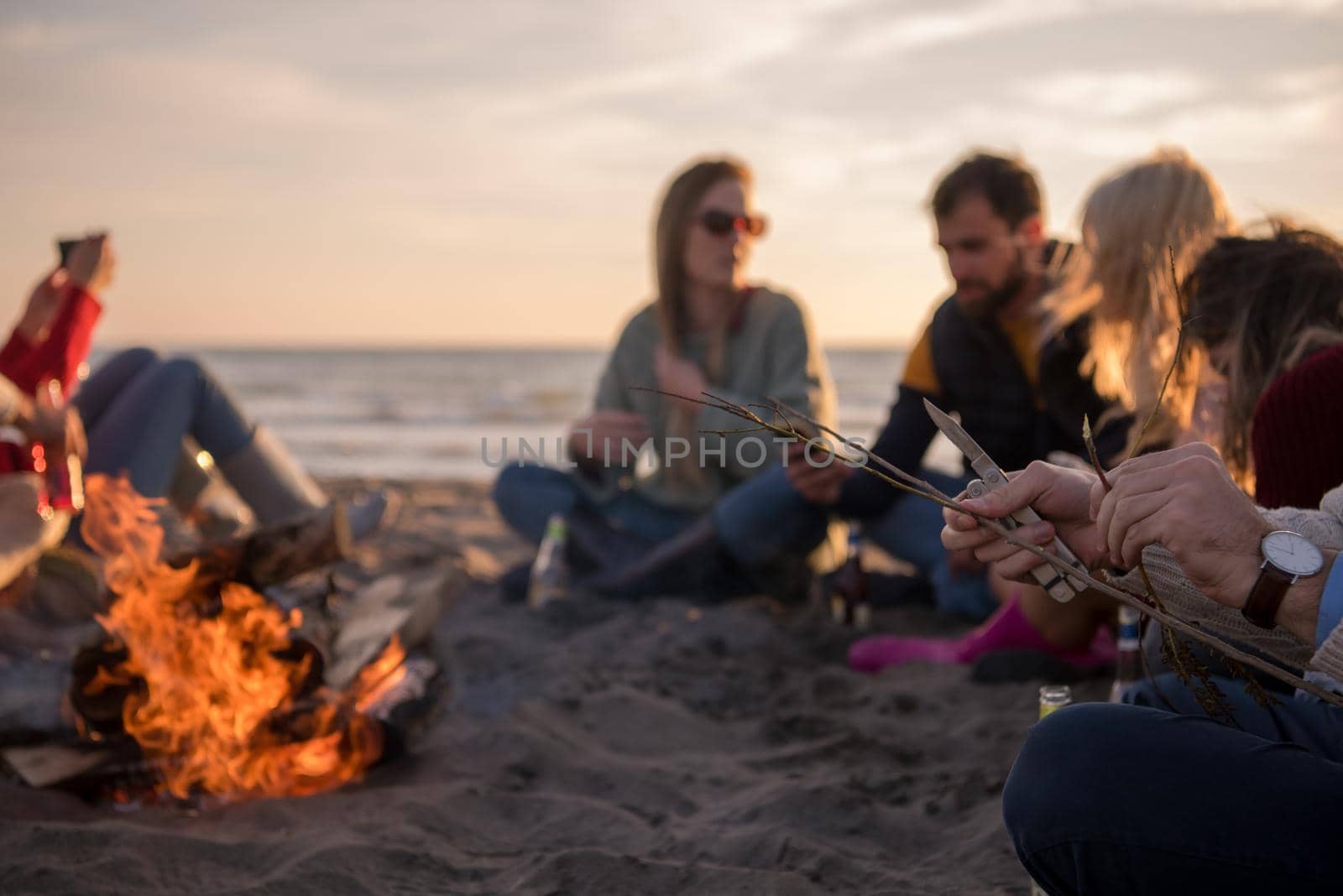 Happy Carefree Young Friends Having Fun And Drinking Beer By Bonefire On The Beach As The Sun Begins To Set