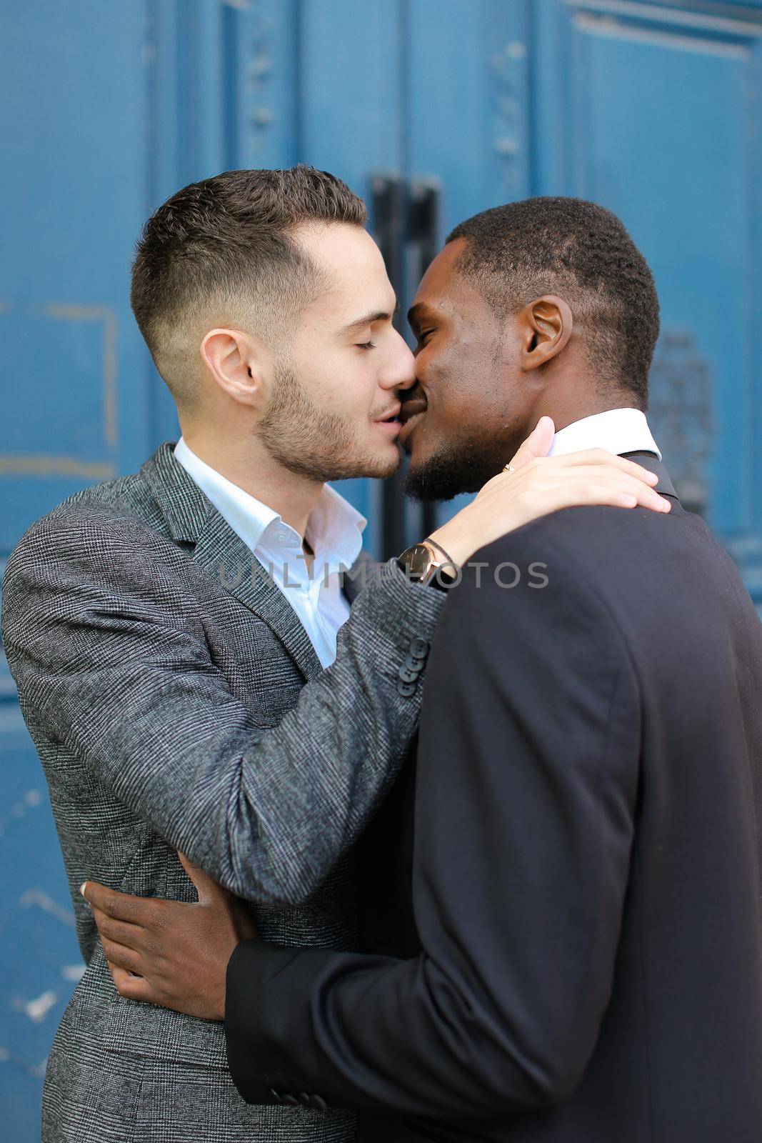 Caucasian happy man kissing afro american guy in door background. Concept of lgbt, gays and same sex couple.