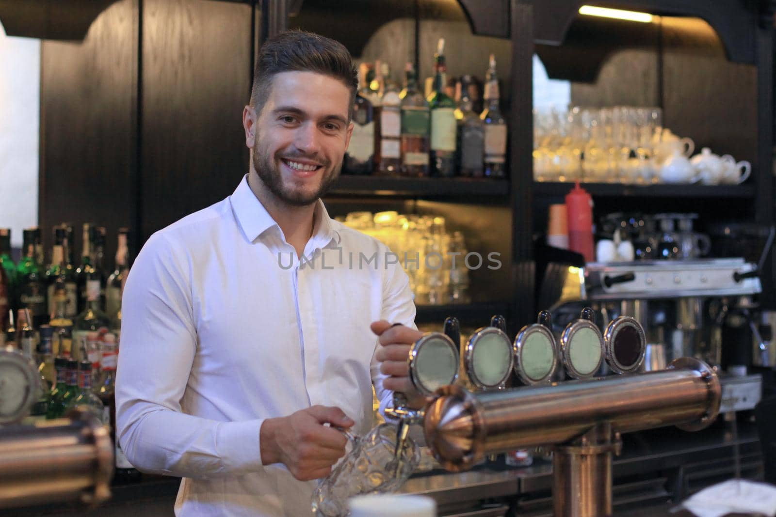 Handsome bartender is smiling and filling a glass with beer while standing at bar counter in pub. by tsyhun