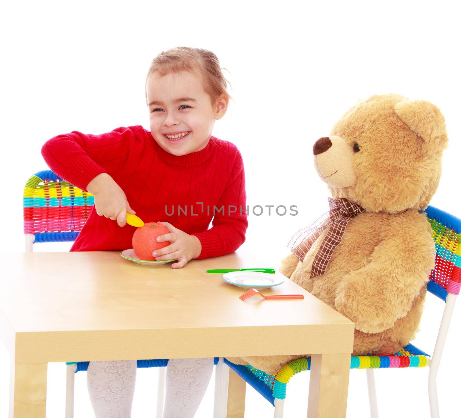 Cute little girl in a red dress in a Montessori kindergarten. Girl sitting at the table with a large Teddy bear and feeds him an Apple - Isolated on white background