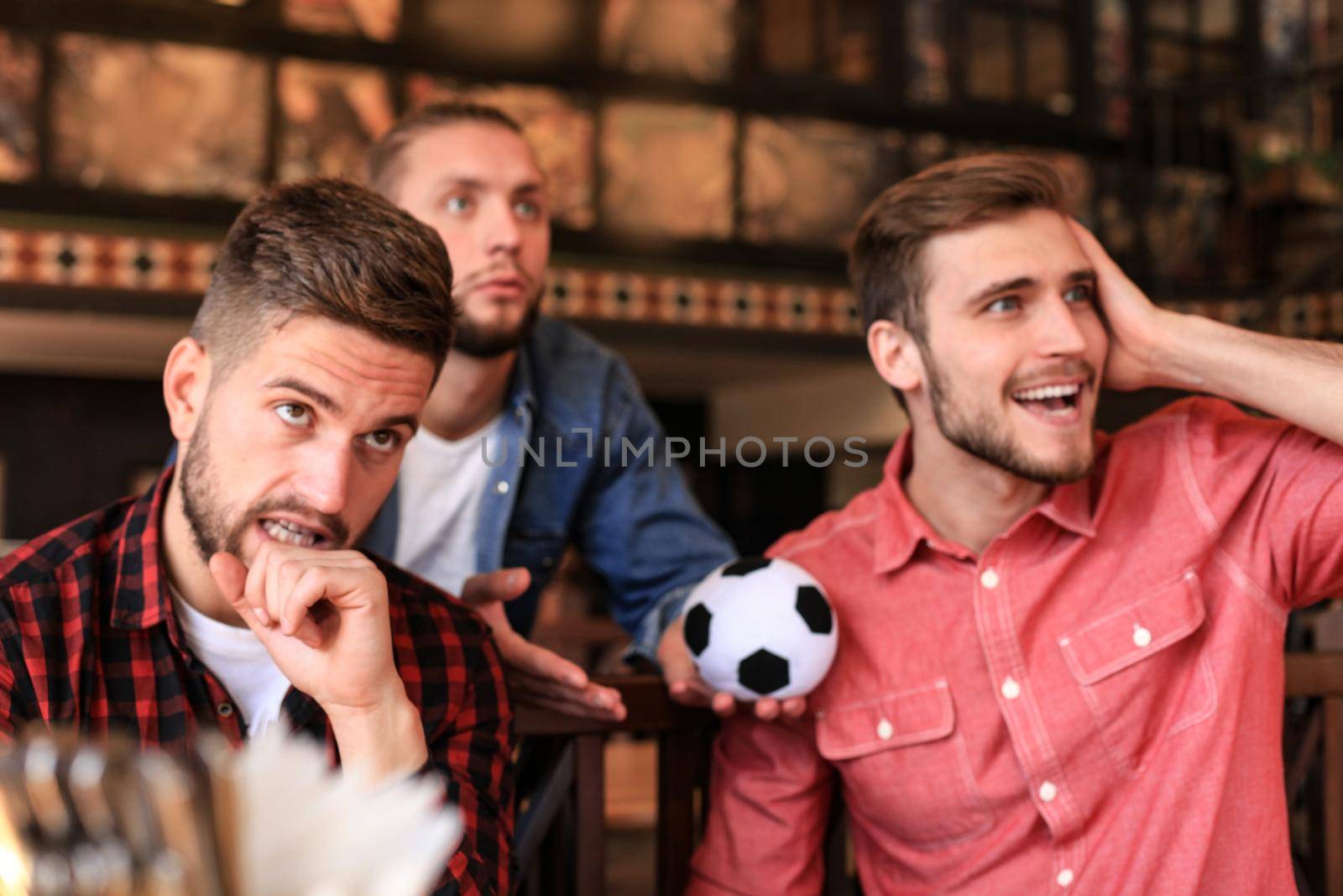 Three men in casual clothes are cheering for football and holding bottles of beer while sitting at bar counter in pub