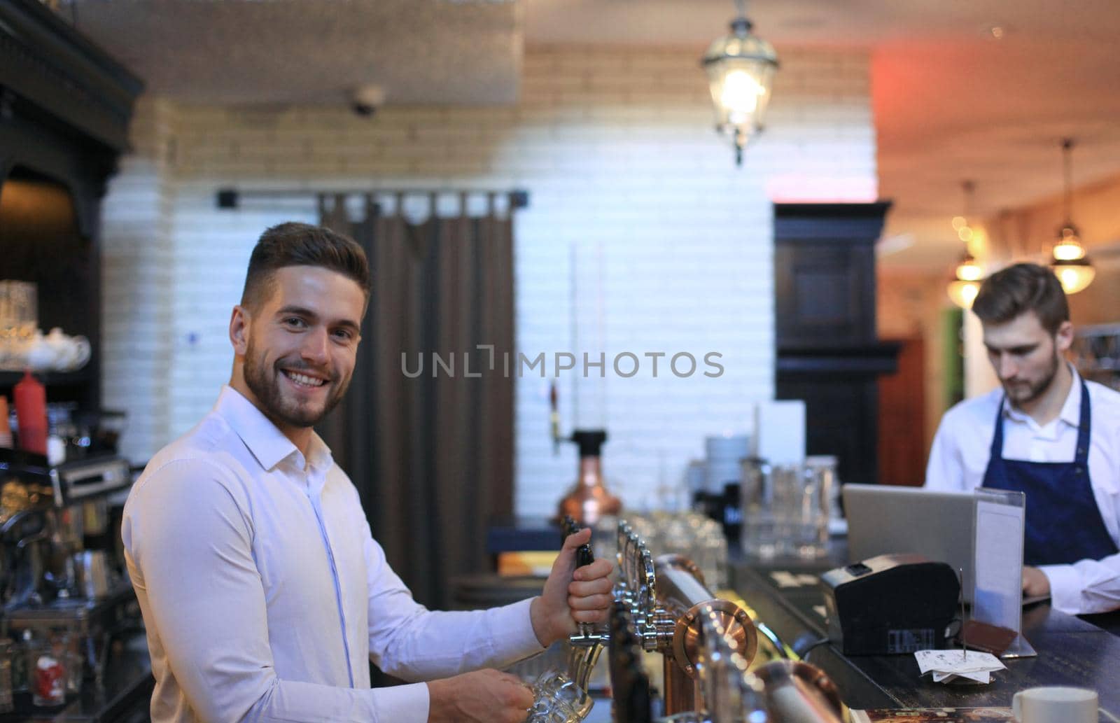 Handsome bartender is smiling and filling a glass with beer while standing at bar counter in pub
