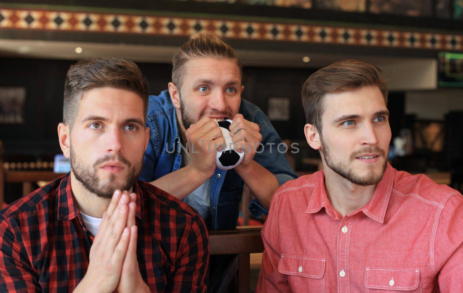 Three men in casual clothes are cheering for football and holding bottles of beer while sitting at bar counter in pub