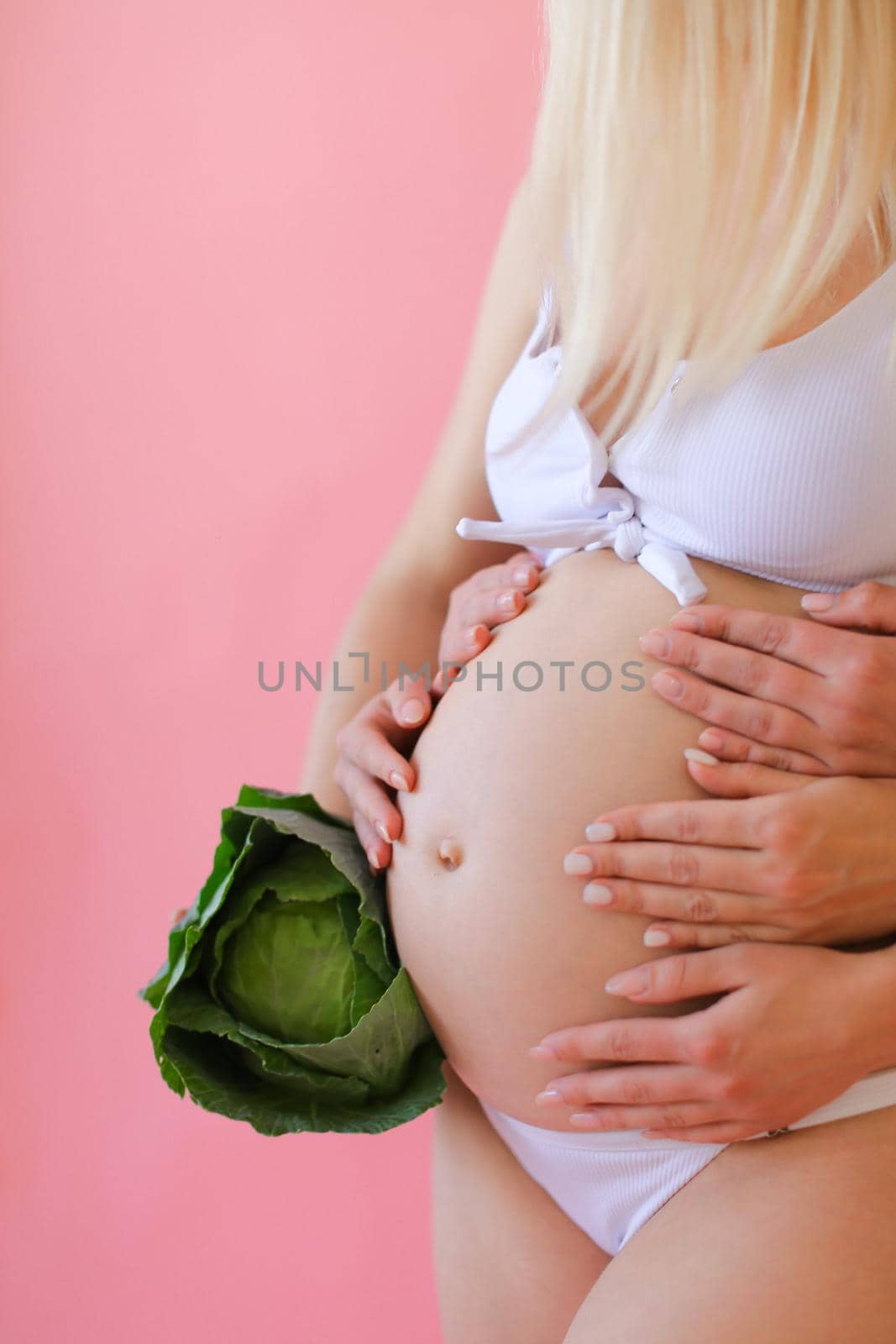 Blonde pregnant woman in underwear holding belly and keeping cabbage in pink monophonic background. Concept of expactant photo session.