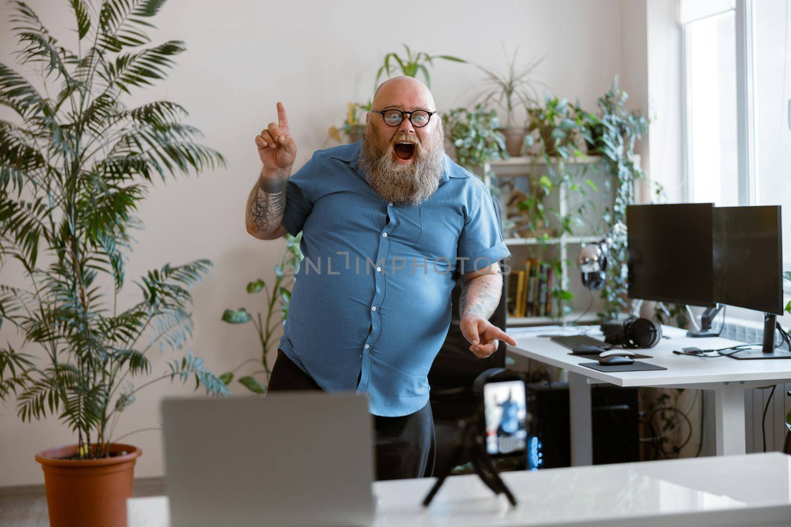 Cheerful funny bearded obese man in tight blue shirt dances recording new video for blog with mobile phone at home