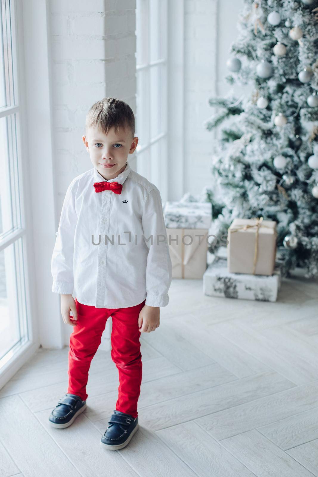 little caucasian boy sits near the christmas tree with a gift by StudioLucky