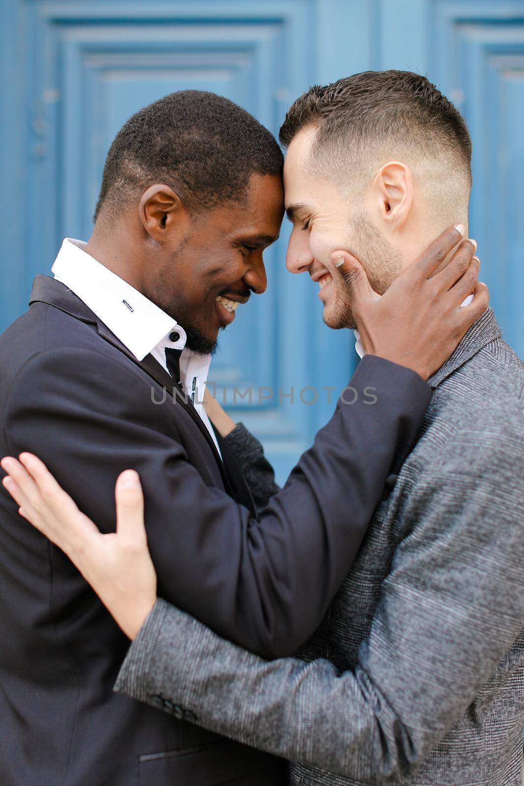 Afro american man hugging caucasian guy in door background, wearing suit. Concept of same sex couple and gays.