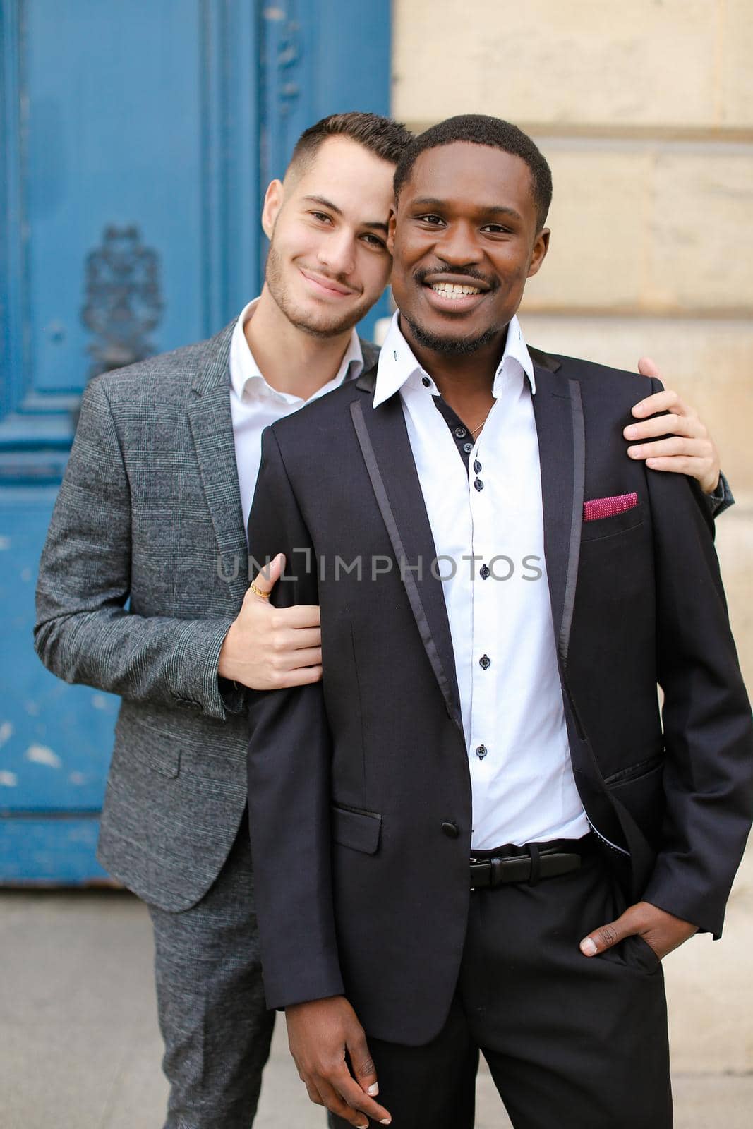 Two smiling men, caucasian and afro american, wearing suits standing near building and hugging. by sisterspro