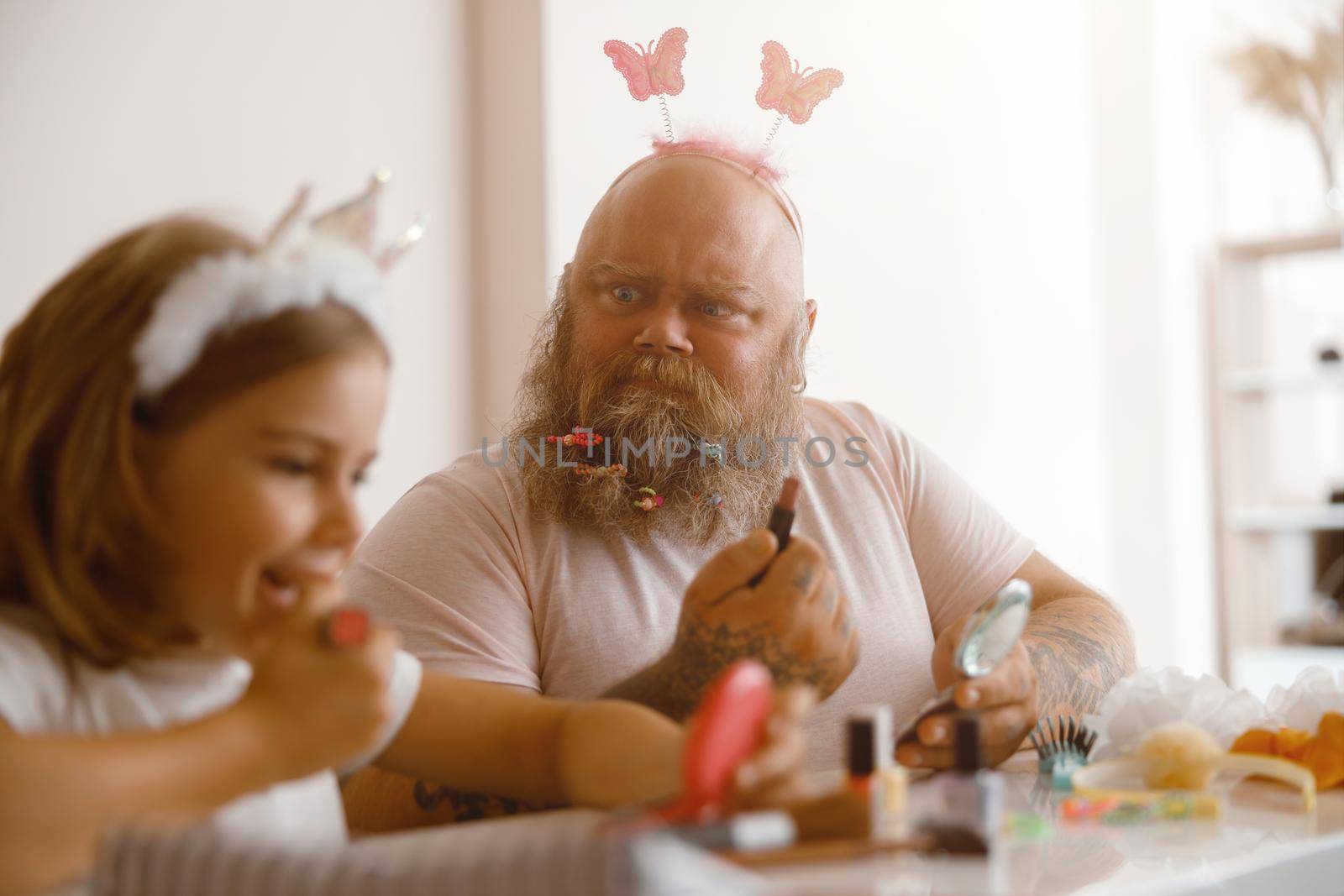 Emotional bearded man with butterflies on headband looks at adorable little girl applying colorful lipstick at table in living room. Beauty salon at home