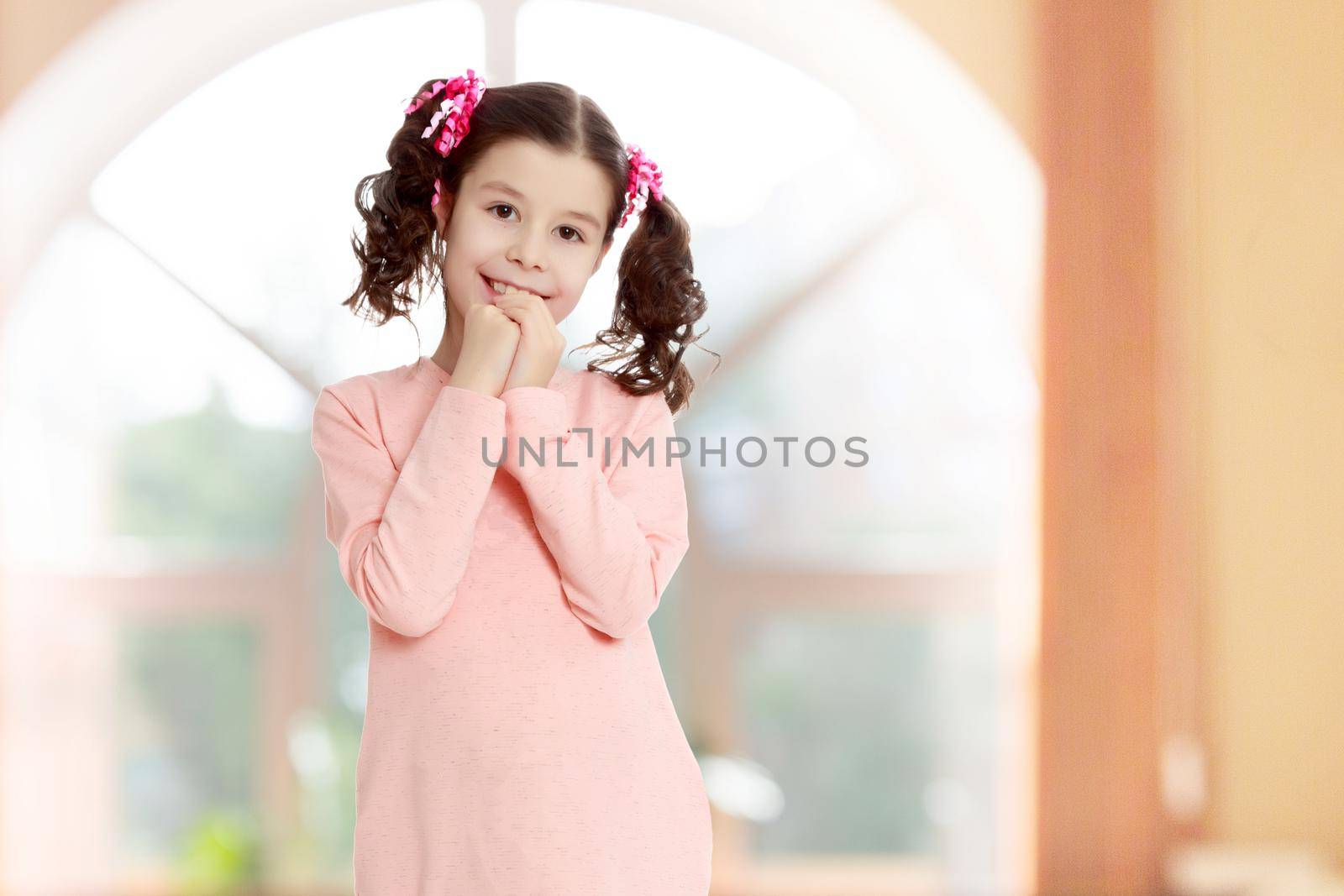 Beautiful little girl with long curly tails on the head,in which braided red ribbons . In long pink dress. The girl shyly holding hands near the face. In a room with a large semi-circular window.