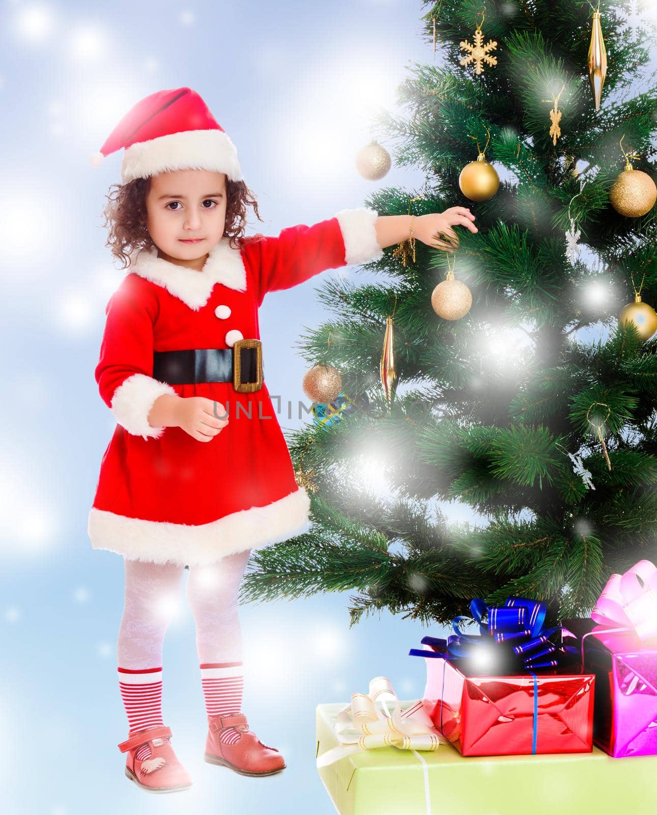 Adorable little curly-haired girl, dressed as Santa Claus decorates a Christmas tree toys.Blue winter background with white snowflakes.