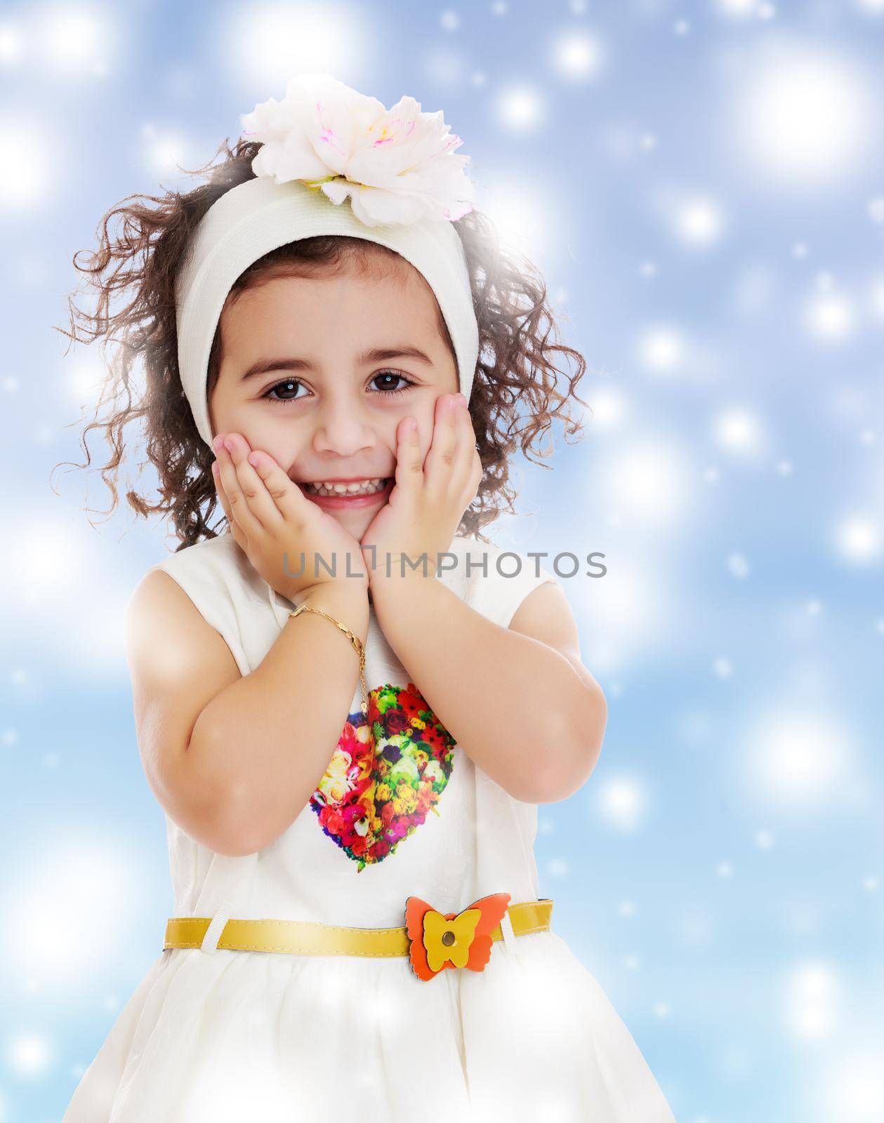 Beautiful little curly girl in a white dress happily surprised. Girl holding hands near the face. Close-up.Blue winter background with white snowflakes.