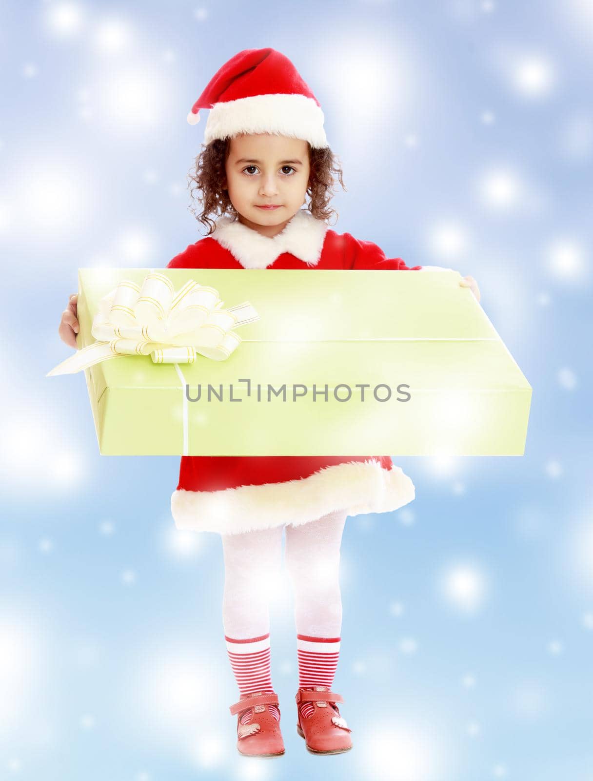 Cute little girl in a coat and hat of Santa Claus, holding a big green box , tied with a bow.Blue winter background with white snowflakes.