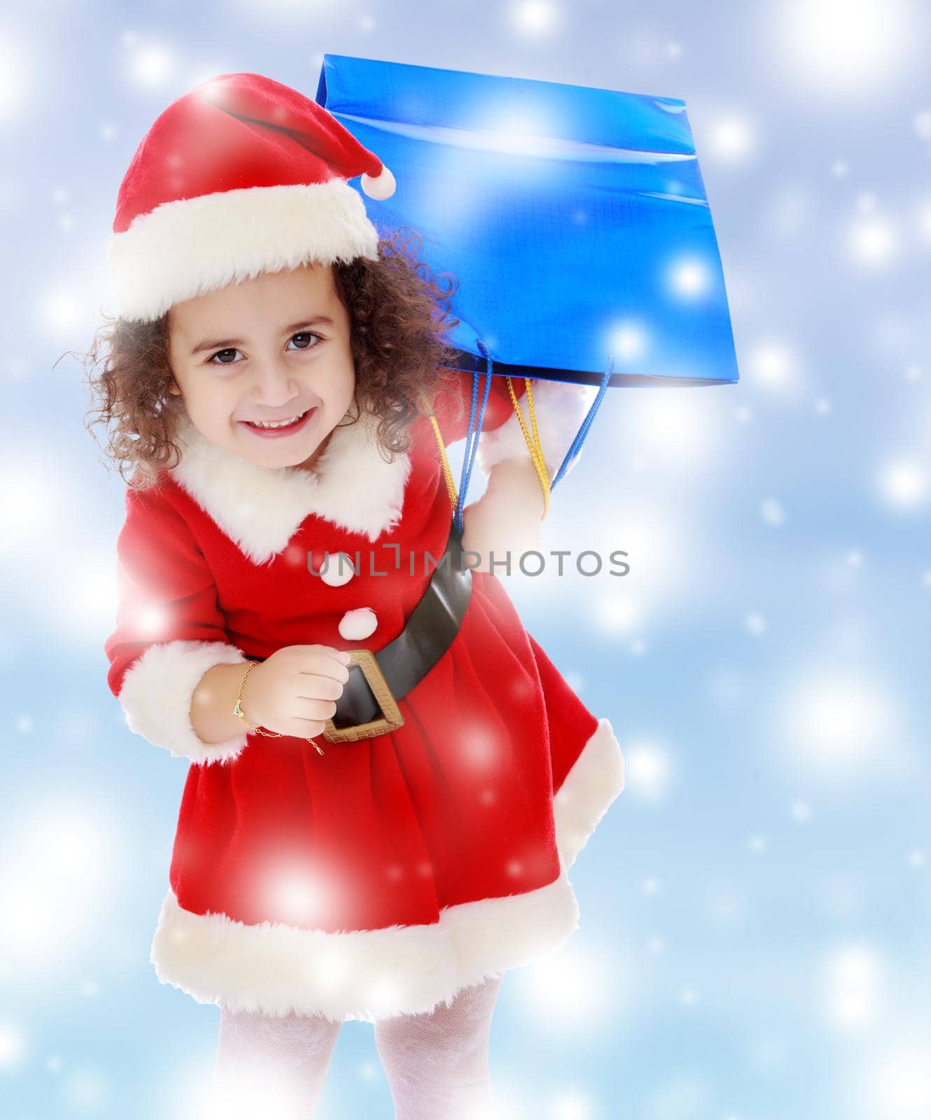 Laughing a little, curly-haired girl in a coat and hat of Santa Claus carries on his shoulder a colorful shopping bags. Close-up.Blue winter background with white snowflakes.