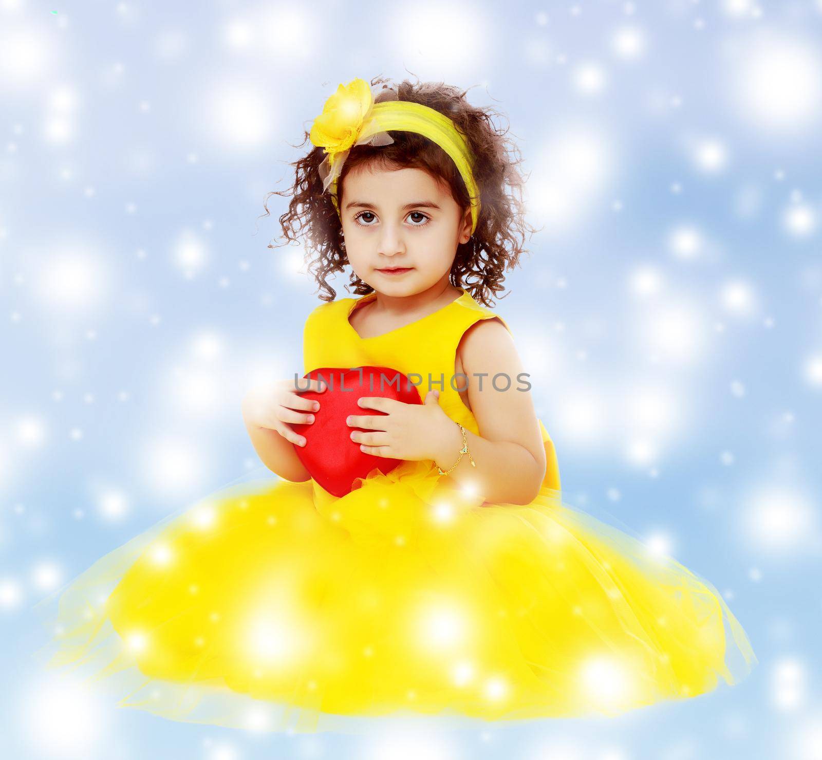 Happy little curly girl in a bright yellow elegant dress sitting on the floor. Girl holding in hand heart.Blue winter background with white snowflakes.