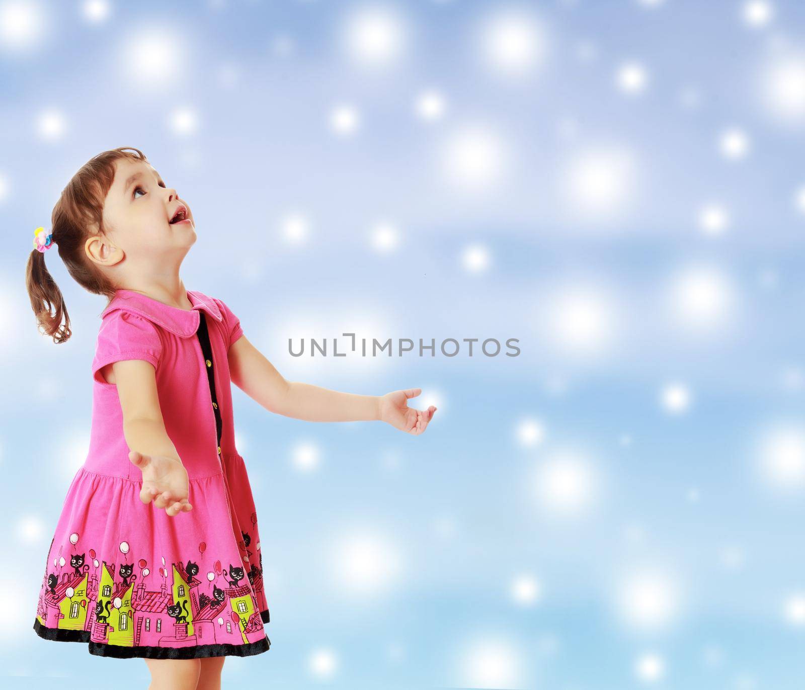 Adorable little girl with pigtails on the head , in a pink dress. The girl was looking at the top turned sideways to the camera.On new year or Christmas blue background with white big stars.