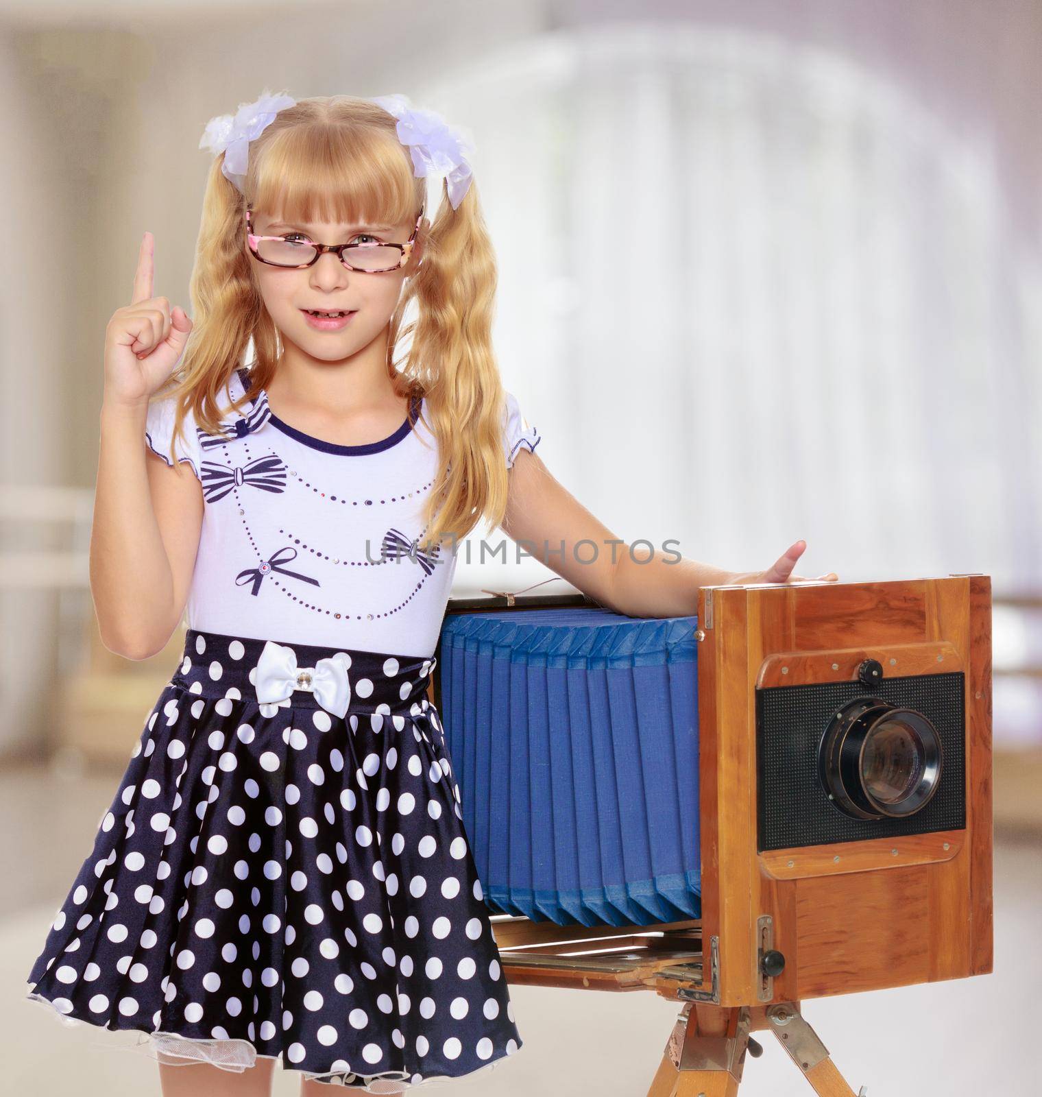Adorable little blond girl wearing glasses and fancy dress polka dot advertises the old wooden camera.In a room with a large semi-circular window.