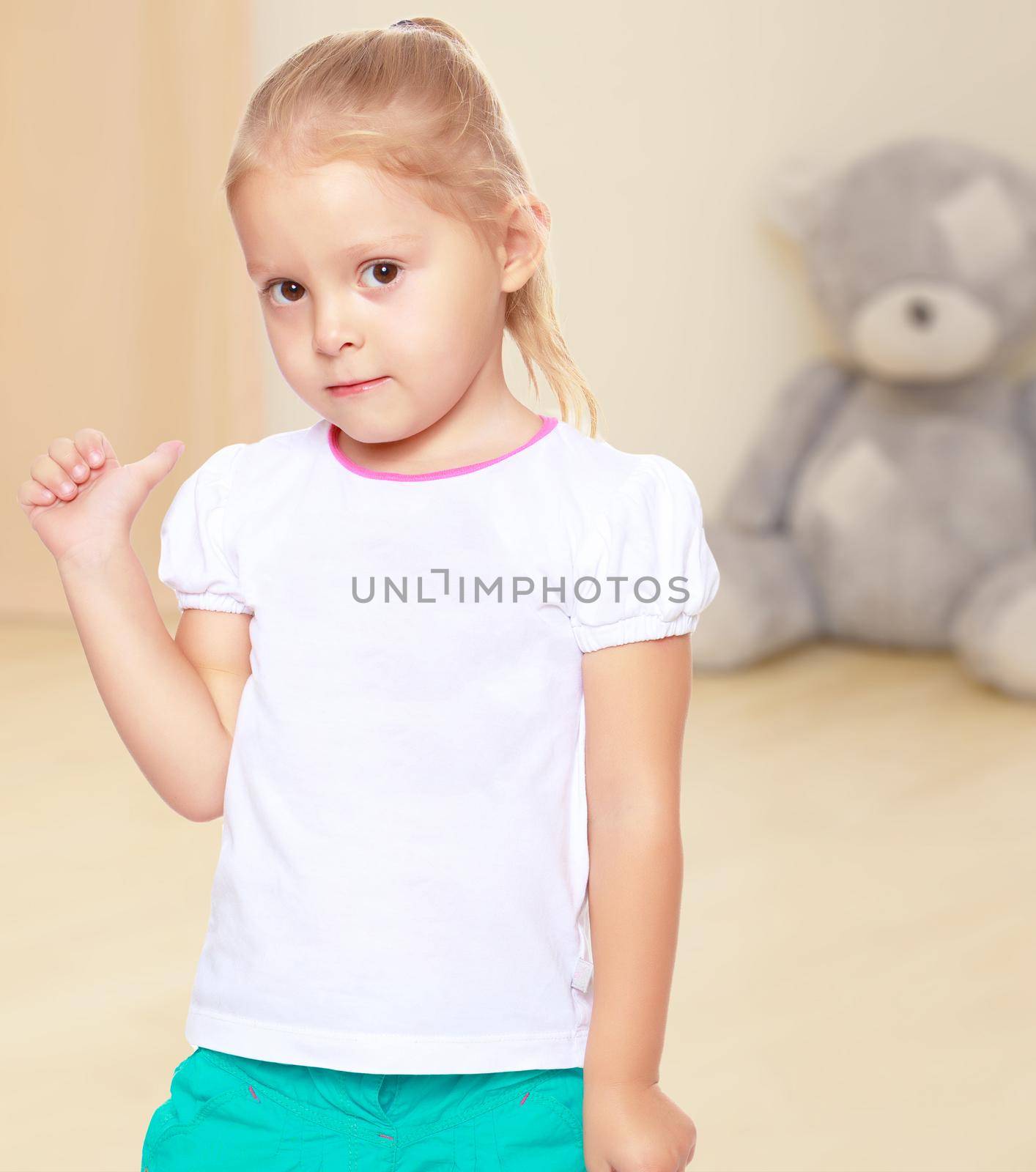 Cute little blond girl in white tank top without a pattern.Girl shows thumb to the side.The concept of preschool development of the child ,against a child's room where in the background a Teddy bear