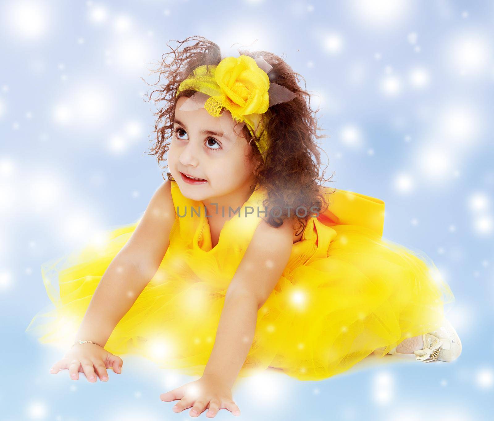 Funny curly little girl in a bright yellow elegant dress crawling on the floor. She looks to the top.Blue winter background with white snowflakes.