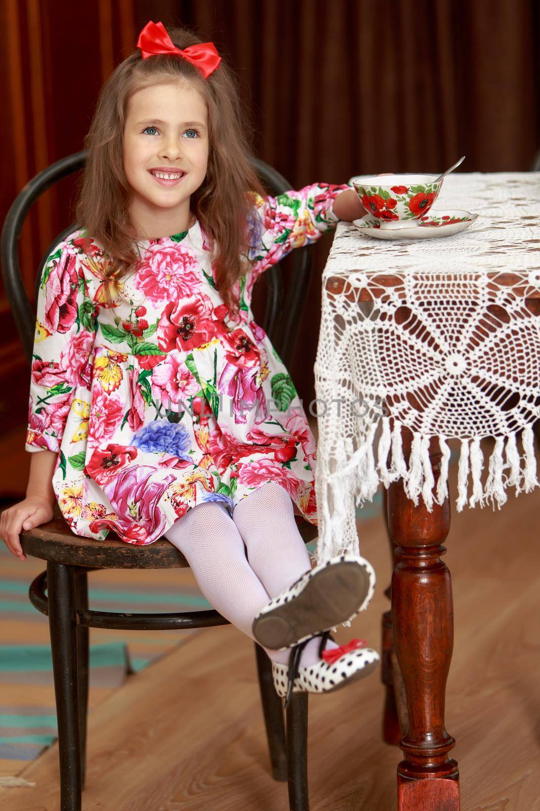 Gentle little girl with a long ponytail down to his shoulders, and a red bow on her head, in a beautiful summer dress. covered with a lace crocheted tablecloth. Beside her is a Cup of tea