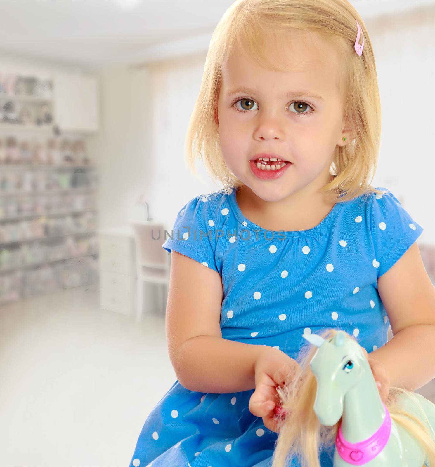 Cute little blonde girl playing with a toy horse. Girl wearing a blue dress with polka dots.On the background of the room where children live, and are on the shelves of toys.