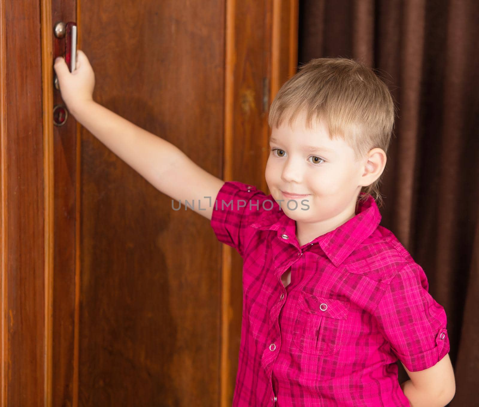 Cute little blond boy in red shirt is posing near the old wardrobe.Retro style, surrounded by old things fifties of the last century.