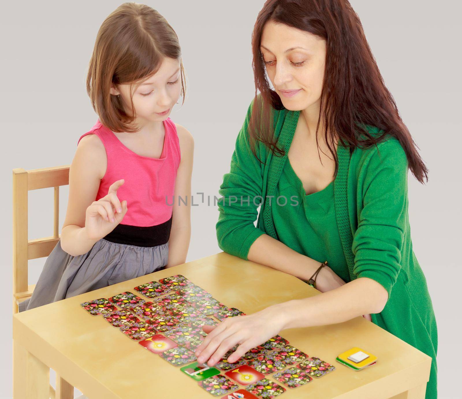 Pensive little girl and her teacher at the table laid out cards with pictures.On a gray background.