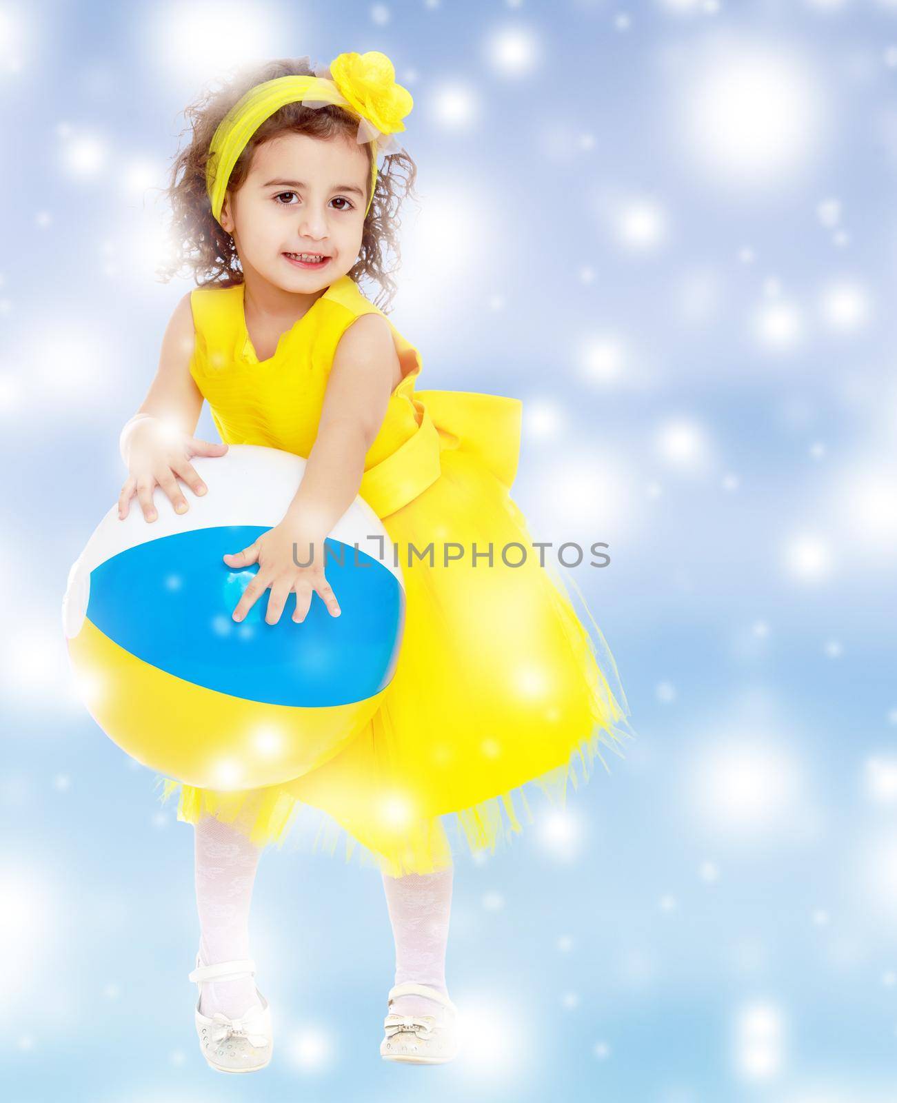 Funny curly baby girl in a bright yellow dress and bow on her head playing with a ball.Blue winter background with white snowflakes.