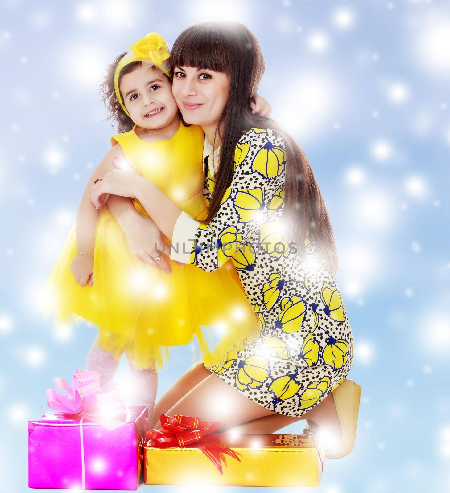 Beautiful young mother and her cute little daughter , embracing, surrounded by Christmas gifts.Blue winter background with white snowflakes.