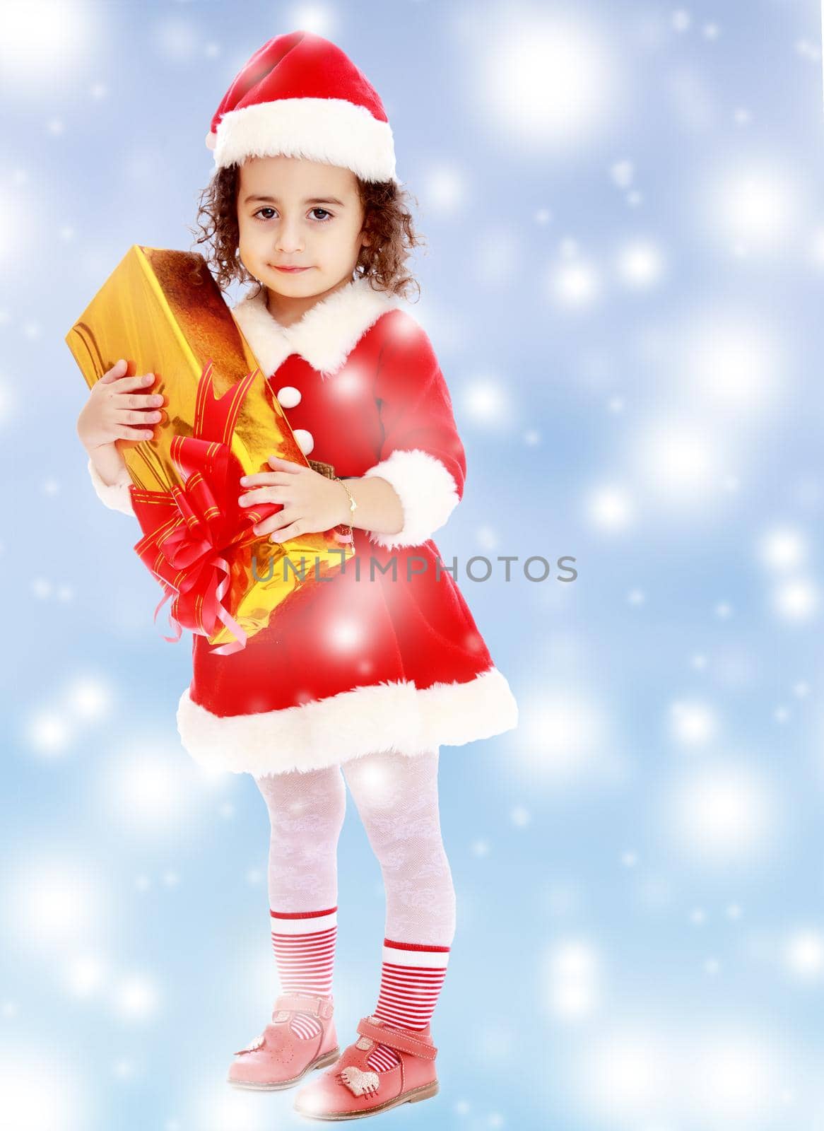 Cute little curly girl in a coat and hat of Santa Claus holding a box with a gift.Blue winter background with white snowflakes.