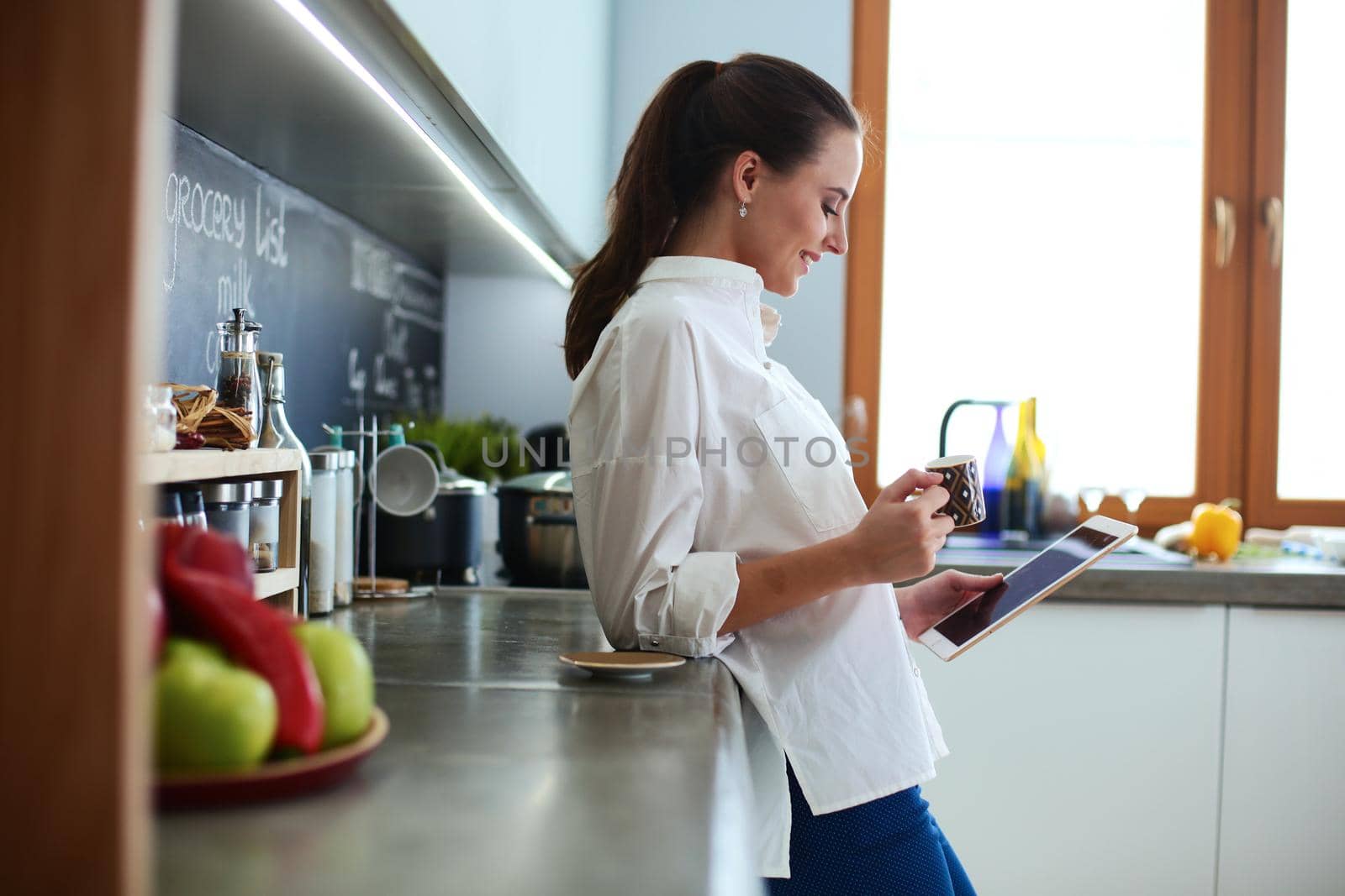 Young woman using tablet in kitchen at home and drinking coffee.