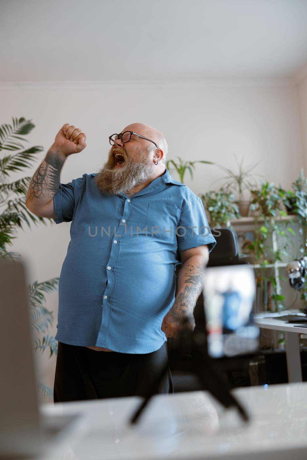 Expressive bearded man with overweight in tight blue shirt sings song recording new video for blog with smartphone at home
