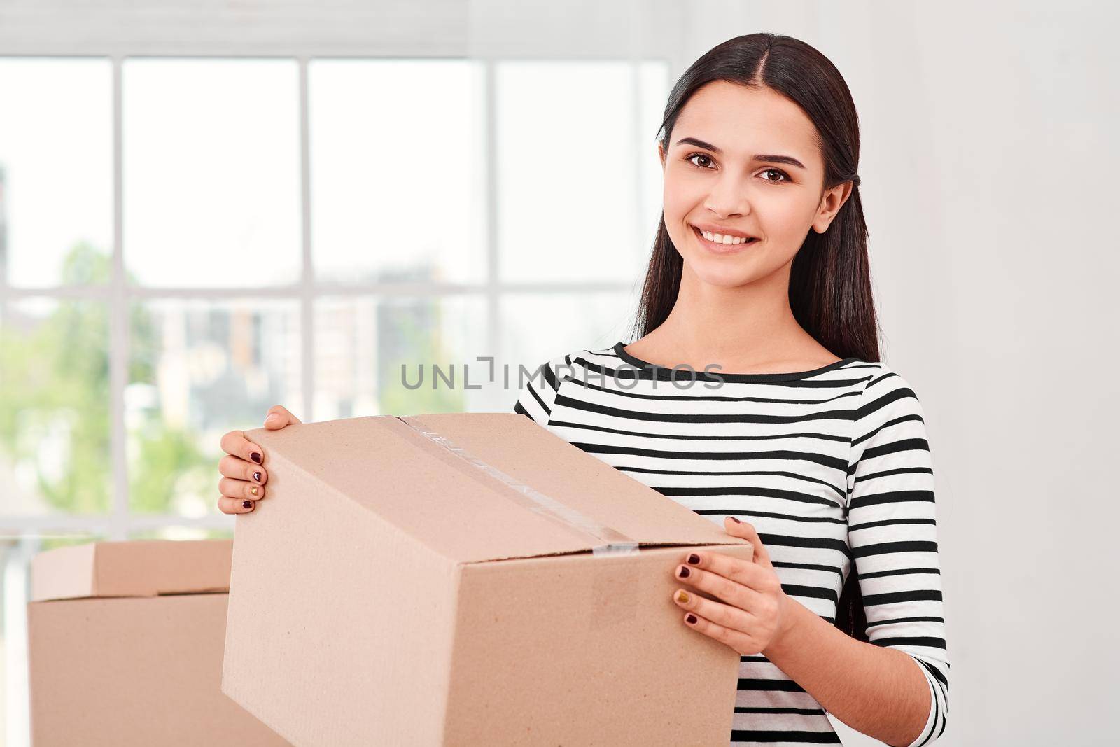 Young entrepreneur, SME, freelance woman working online with clients making purchases, orders. She is preparing packages. Woman standing near boxes and looking at the camera with a smile. Concept of success