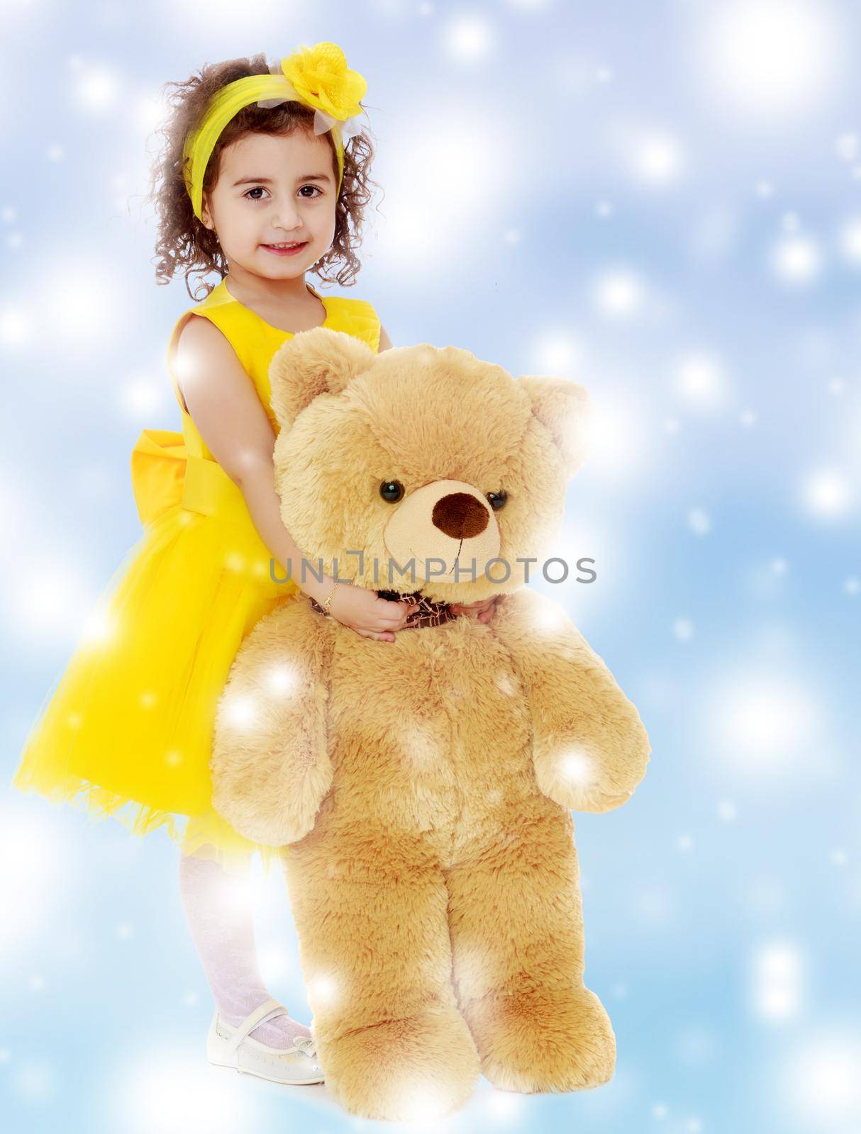 Joyful little girl Princess, dressed in a yellow dress, hugging a big Teddy bear. Standing at full height.Blue winter background with white snowflakes.