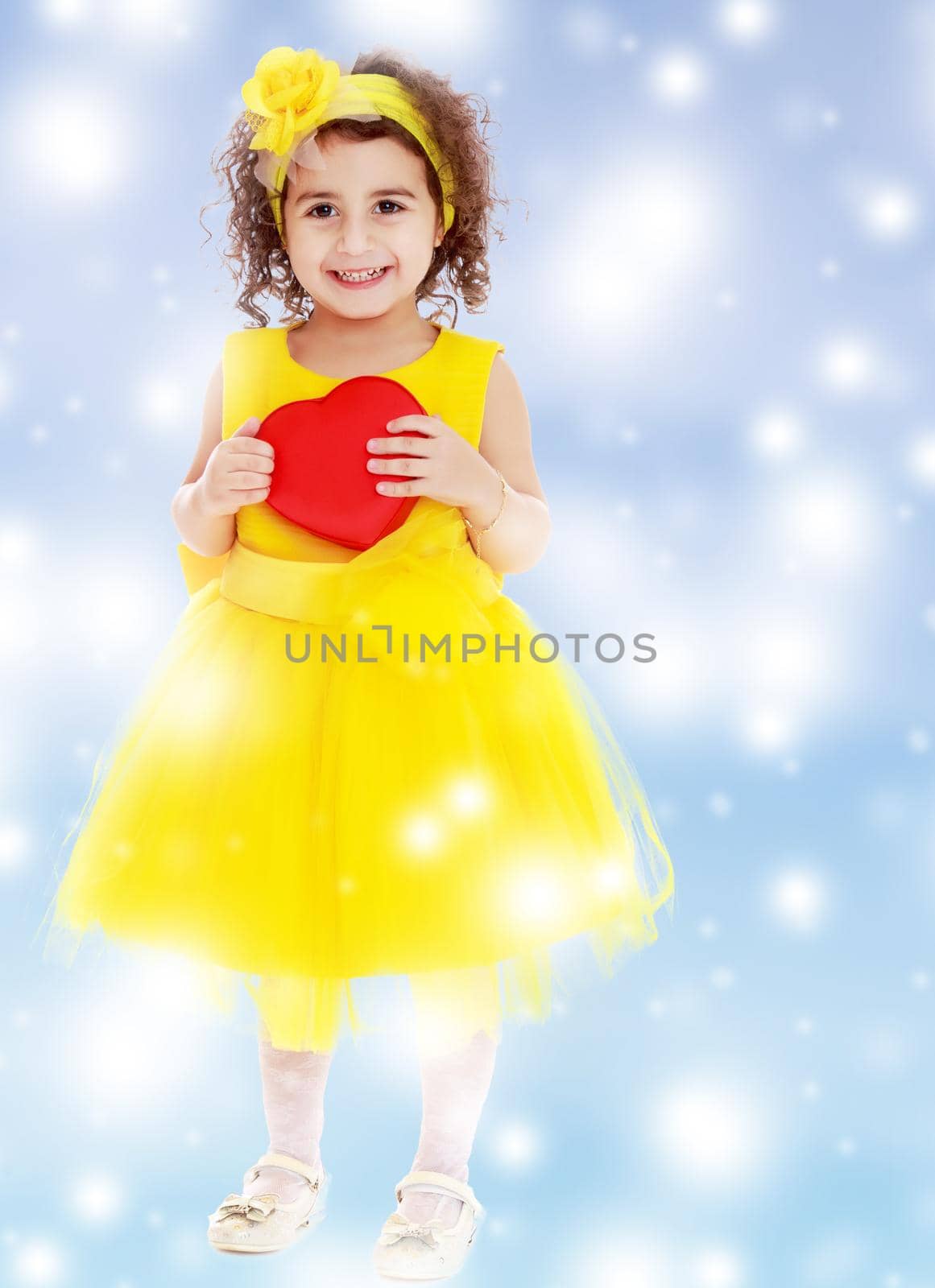 Adorable little girl in bright yellow elegant dress. Close to her heart.Blue winter background with white snowflakes.