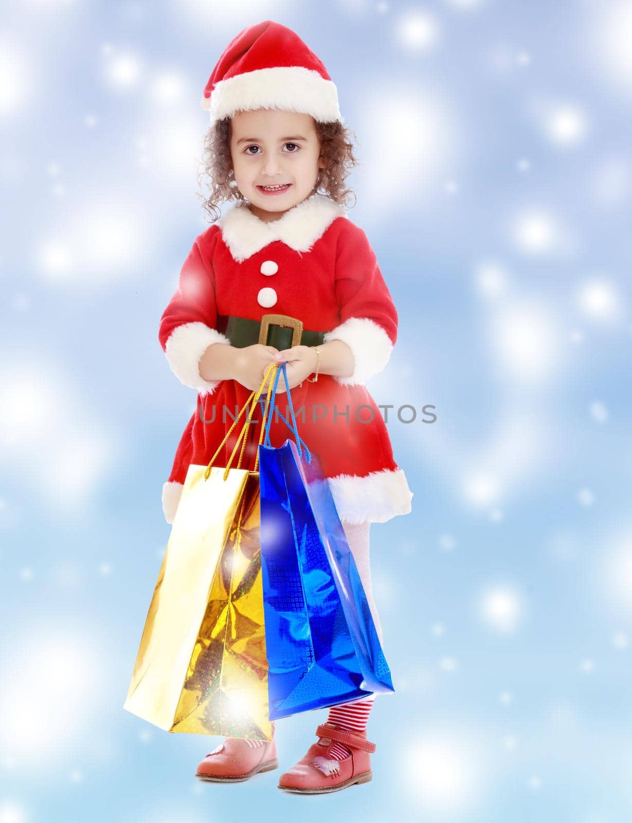 Adorable little curly-haired girl in a coat and hat of Santa Claus,holding colorful shopping bags.Blue winter background with white snowflakes.