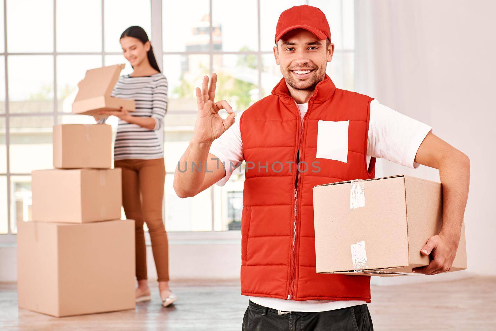 Young courier in red postal uniform standing with parcel indoors, showing OK sign, looking at the camera with a smile. He has done his job very well. Woman unpacking her purchases in the background. A stack of carton boxes. Bright interior. Friendly worker, high quality delivery service