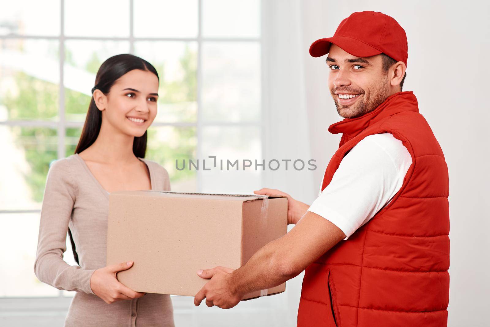 Cheerful postman wearing red postal uniform is delivering parcel to a satisfied client. He is looking at the camera, while she is looking at him. They both hold carton box and smiling. Friendly worker, high quality delivery service. Indoors. Bright interior.
