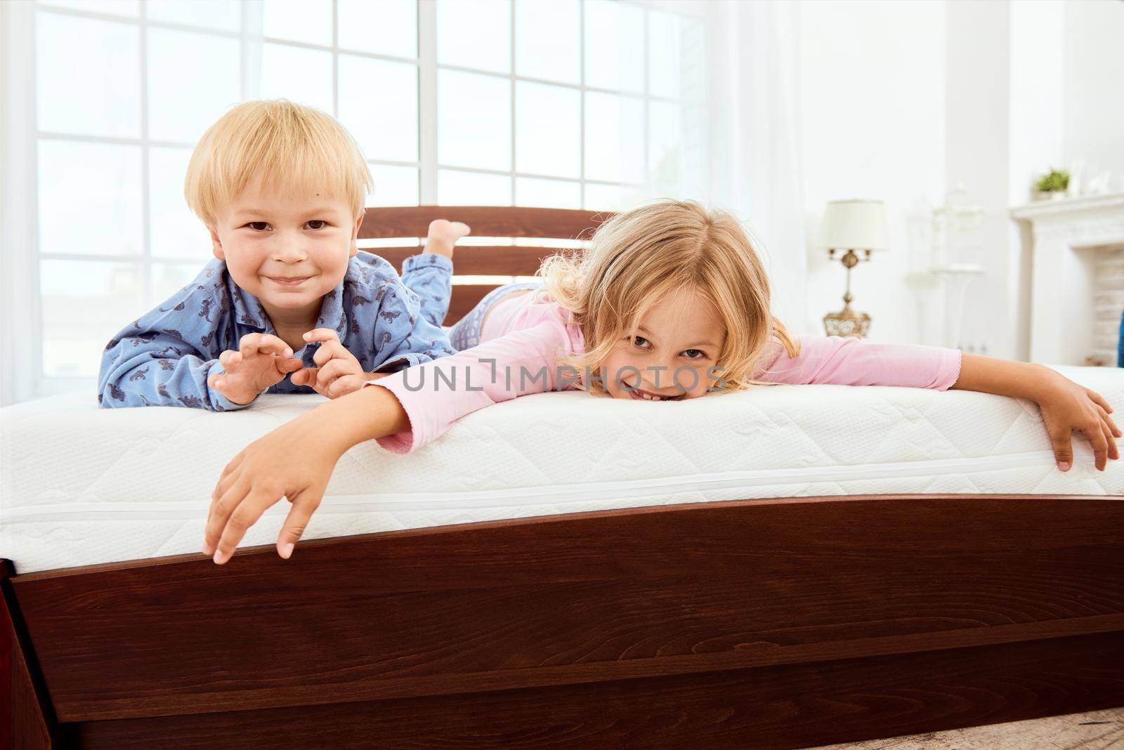 Happy cute little brother and sister looking at camera and smiling while lying on a large comfortable white mattress in bedroom. Playing together. Quarantine. Bedroom. Family