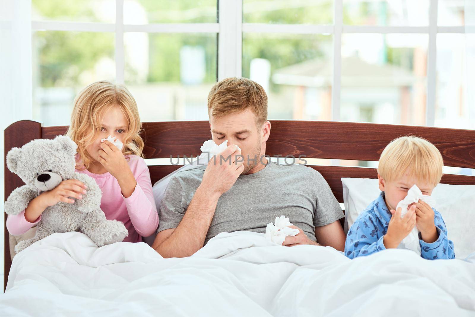 Young father and his children suffering from flu or cold and wiping their noses while lying in bed together. Virus disease. Coronavirus concept. Sick family at home. Health concept