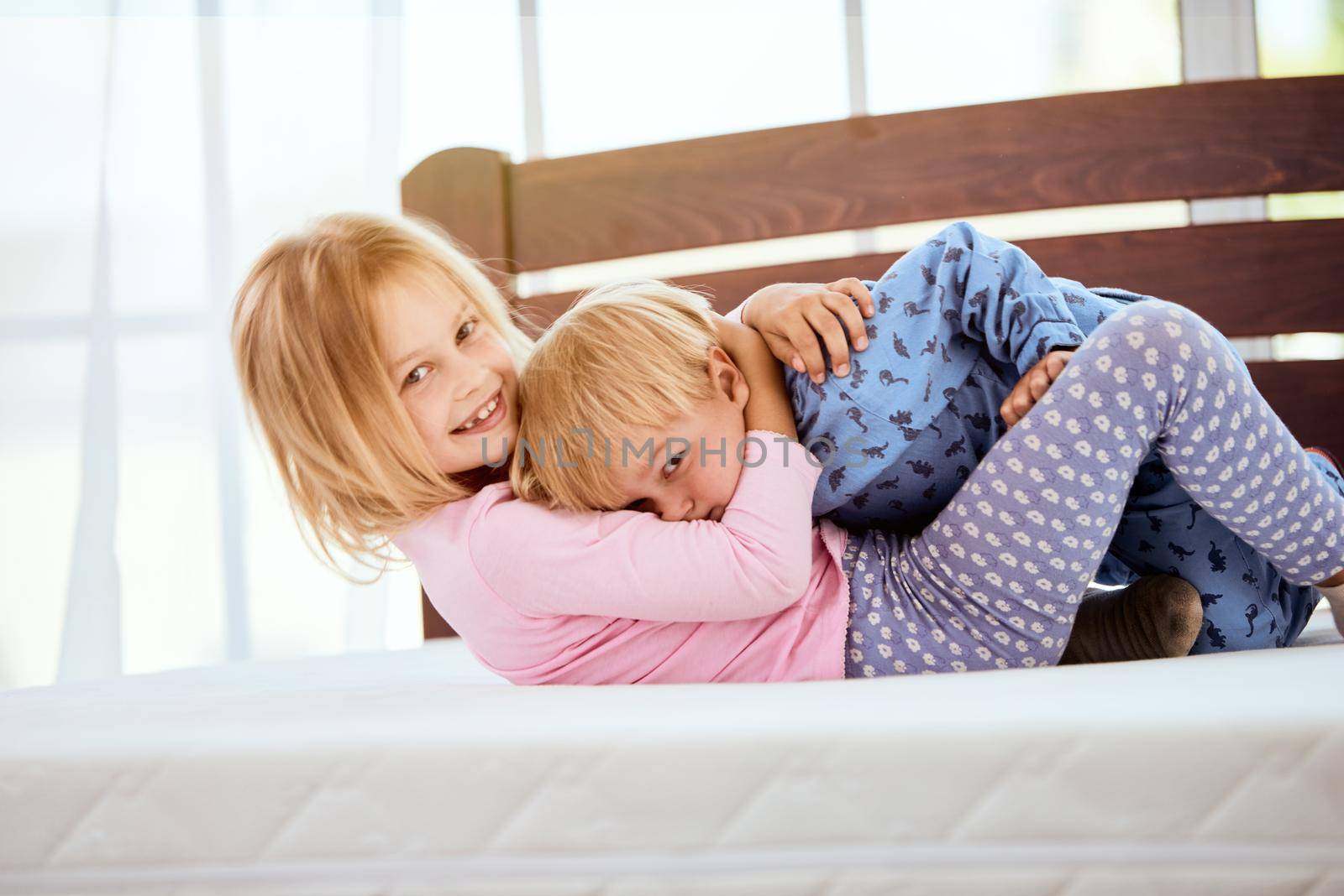 Happy little sister and brother in pajamas playing and having fun while lying on a large comfortable white mattress in bedroom. Happy family at home