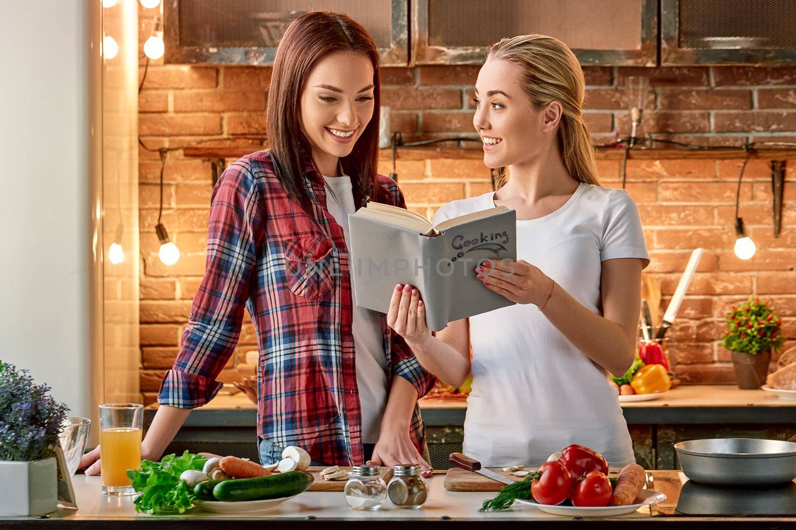 Portrait of two attractive women, having fun, while preparing salad. They are fully involved in the process. Blonde girl in white T-shirt and her dark-haired friend in checkered shirt searching for healthy recipes in a book. They decide what to cook today. Front view