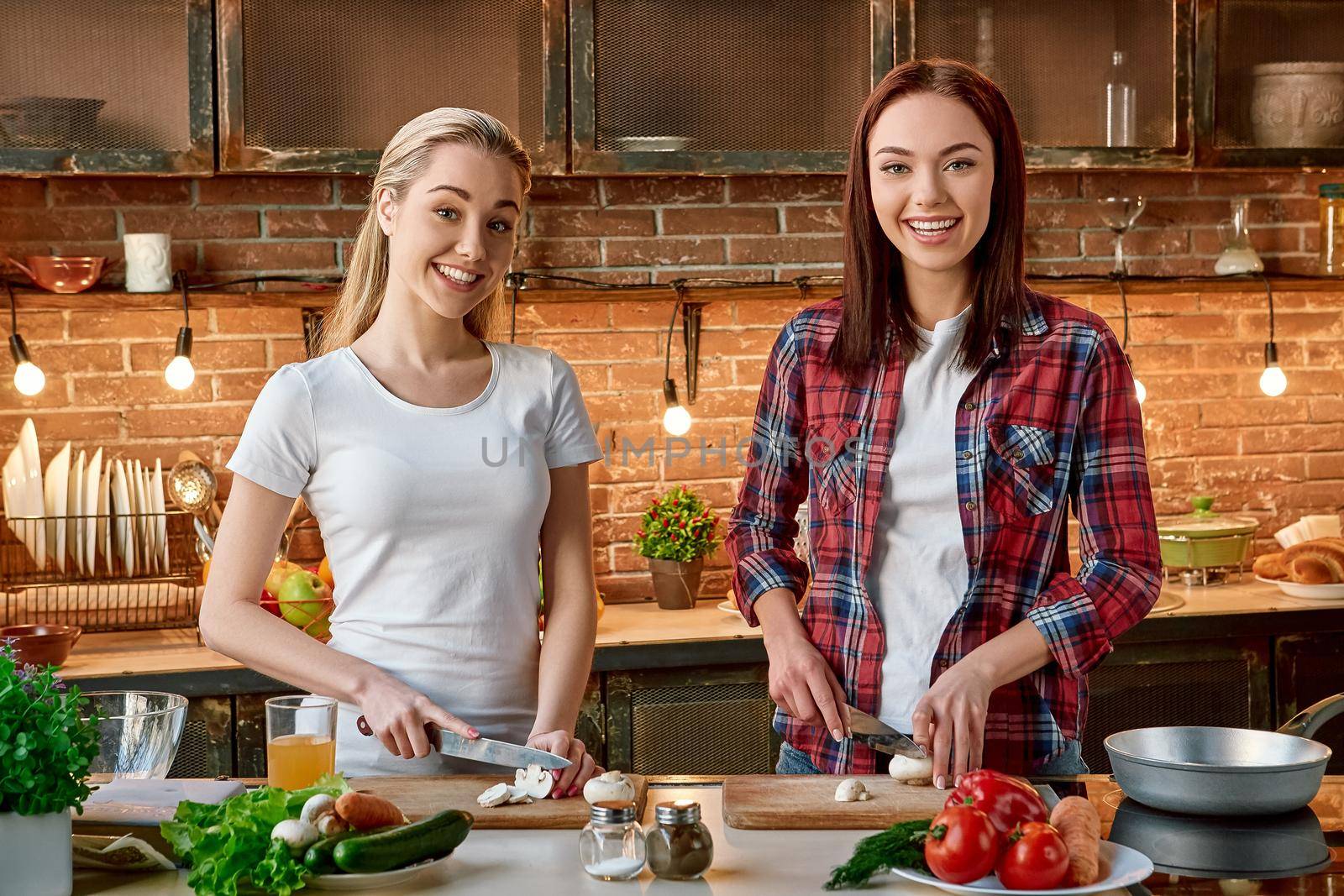 Portrait of two attractive women, having fun, while preparing vegetable salad. They are fully involved in the process. They are looking at the camera and smiling while slicing mushrooms. Front view